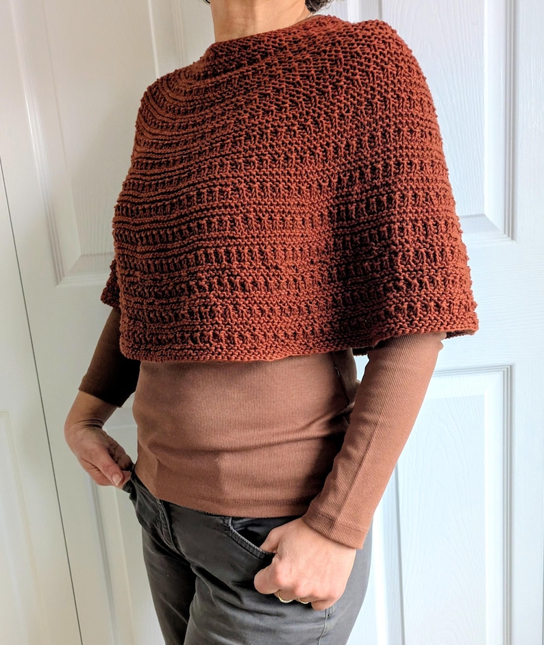 Pode incluir: Um poncho tricotado &agrave; m&atilde;o na cor ferrugem com um padr&atilde;o de malha canelada texturizada. A pe&ccedil;a tem um decote alto e arredondado e uma silhueta curta tipo capa que cai sobre os ombros, usada sobre uma camisa castanha de manga comprida.