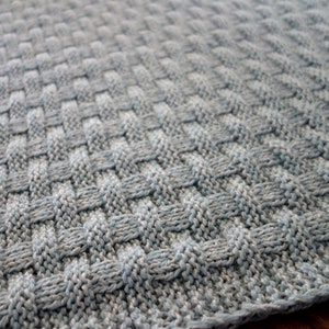 May include: A close-up of a knitted baby blanket in a light blue and gray color scheme. The blanket has a woven texture and is made with soft yarn.
