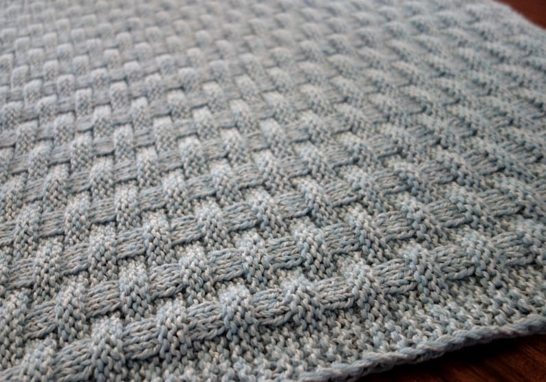 May include: A close-up of a knitted baby blanket in a light blue and gray color scheme. The blanket has a woven texture and is made with soft yarn.