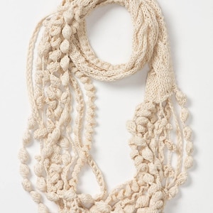 Op de afbeelding: Een crèmekleurige gehaakte sjaal met meerdere strengen van verschillende texturen en pompoenen.