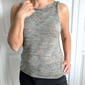May include: A sleeveless knit top in a speckled pattern of blue, brown, and grey. The top has a rounded neckline and a slightly curved hem. The fabric appears to be a soft, textured yarn. The person is wearing black pants.