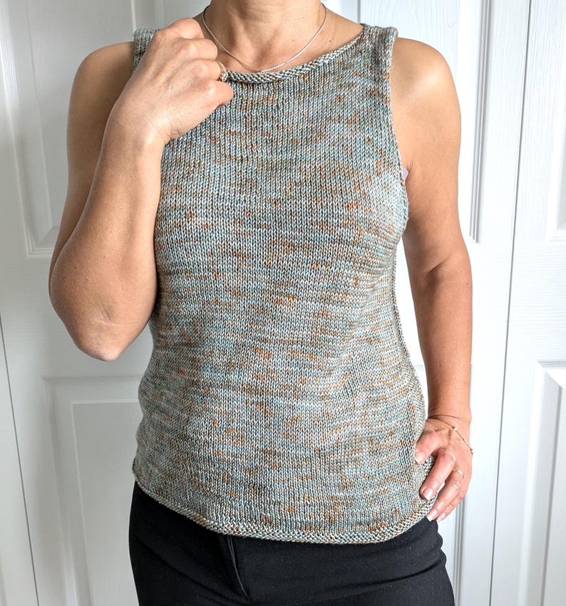 May include: A sleeveless knit top in a speckled pattern of blue, brown, and grey. The top has a rounded neckline and a slightly curved hem. The fabric appears to be a soft, textured yarn. The person is wearing black pants.