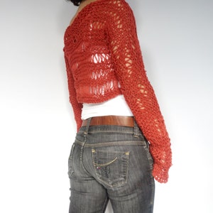 May include: A red crocheted cropped sweater with a loose, open knit pattern. The sweater has long sleeves and a relaxed fit.