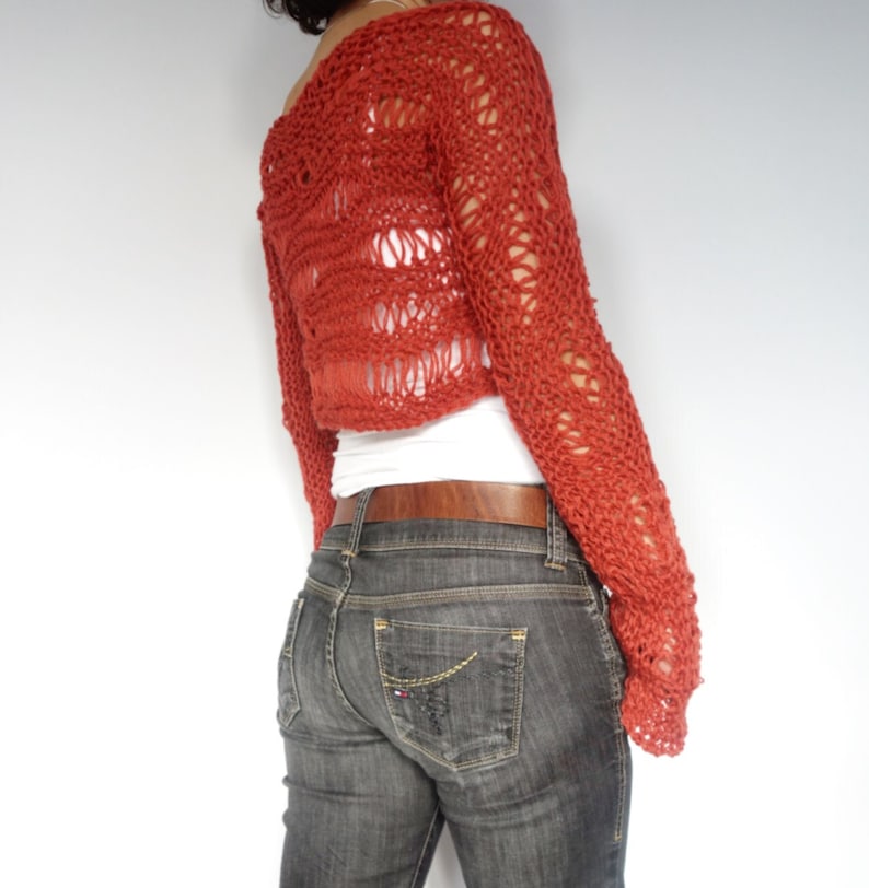 May include: A red crocheted cropped sweater with a loose, open knit pattern. The sweater has long sleeves and a relaxed fit.