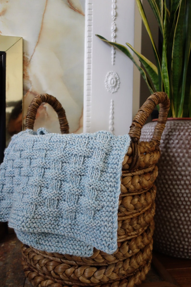 May include: A woven wicker basket with a light blue knitted blanket inside. The basket has two handles and is made of natural materials.