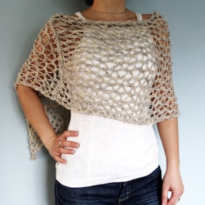 May include: A beige crochet poncho with a delicate openwork pattern. The poncho is worn over a white tank top and blue jeans.