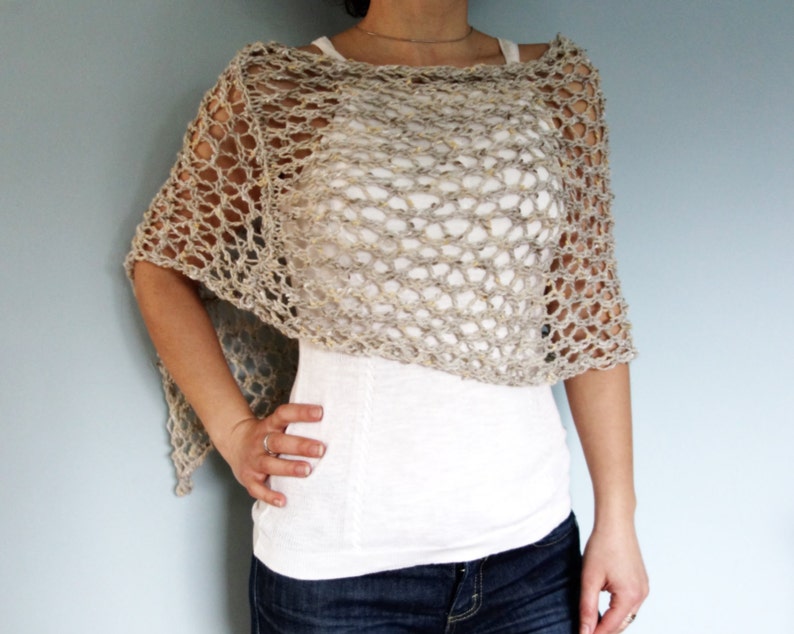 May include: A beige crochet poncho with a delicate openwork pattern. The poncho is worn over a white tank top and blue jeans.
