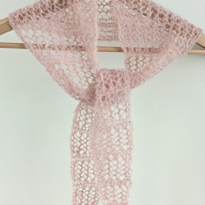 May include: A delicate, light pink scarf with an open, lacy weave. The scarf is displayed on a wooden hanger against a white background. The scarf's texture appears soft and airy, suggesting a lightweight material.