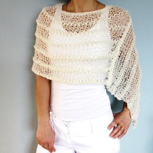 May include: A white crocheted shawl with a delicate openwork pattern. The shawl is draped over the shoulders of a person wearing a white top and white pants.