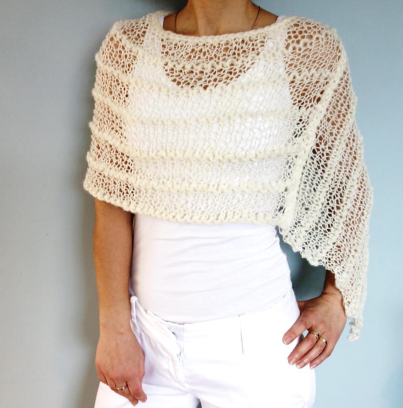 May include: A white crocheted shawl with a delicate openwork pattern. The shawl is draped over the shoulders of a person wearing a white top and white pants.