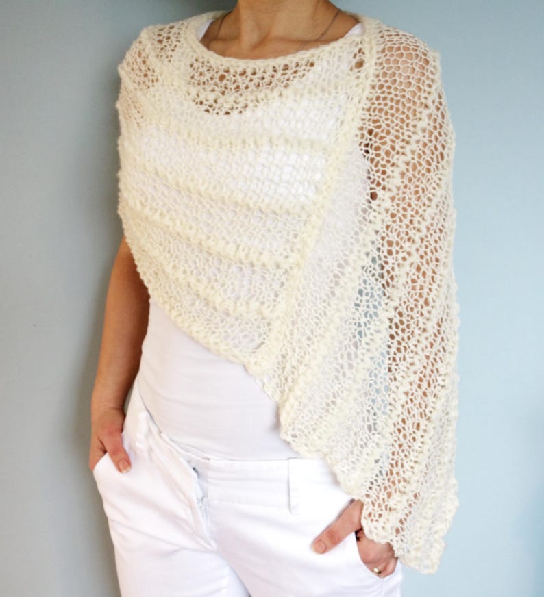 May include: A white crocheted shawl with a delicate openwork pattern. The shawl is draped over the shoulders of a person wearing a white shirt and white pants.