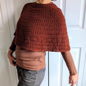 Pode incluir: Um poncho tricotado &agrave; m&atilde;o na cor ferrugem com um padr&atilde;o de pontos texturizados e repetitivos. A pe&ccedil;a tem um decote redondo e uma silhueta curta tipo capa que cai sobre os ombros, usada sobre uma camisa castanha de manga comprida.