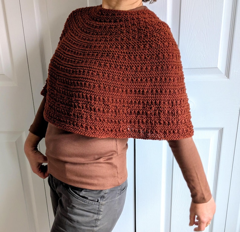 Pode incluir: Um poncho tricotado &agrave; m&atilde;o na cor ferrugem com um padr&atilde;o de pontos texturizados e repetitivos. A pe&ccedil;a tem um decote redondo e uma silhueta curta tipo capa que cai sobre os ombros, usada sobre uma camisa castanha de manga comprida.