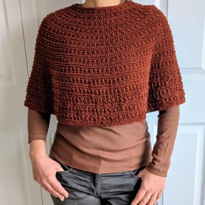 Pode incluir: Um poncho curto de cor ferrugem, tricotado &agrave; m&atilde;o, com um padr&atilde;o de pontos texturizados repetitivos. A pe&ccedil;a apresenta um decote circular largo e mangas curtas e largas que terminam acima dos cotovelos, sobre uma camisola castanha de manga comprida.