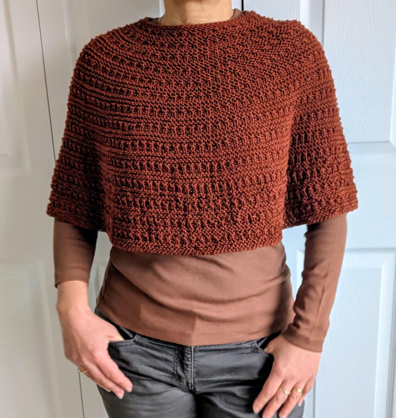 Pode incluir: Um poncho curto de cor ferrugem, tricotado &agrave; m&atilde;o, com um padr&atilde;o de pontos texturizados repetitivos. A pe&ccedil;a apresenta um decote circular largo e mangas curtas e largas que terminam acima dos cotovelos, sobre uma camisola castanha de manga comprida.