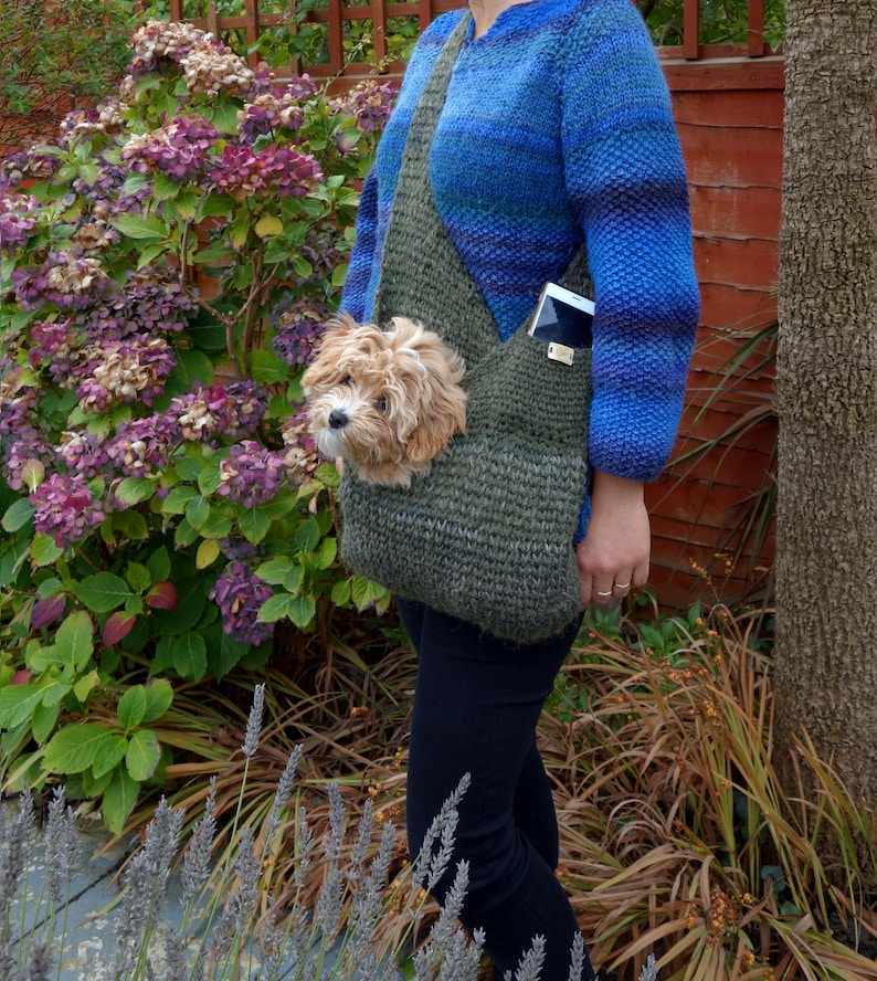 May include: A woman wearing a blue knitted sweater carries a small dog in a green knitted sling bag. The bag has a pocket on the front that holds a cell phone.