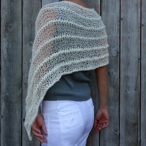May include: A white crocheted shawl with a lacy pattern. The shawl is draped over a person's shoulders and is worn with a gray shirt and white pants.