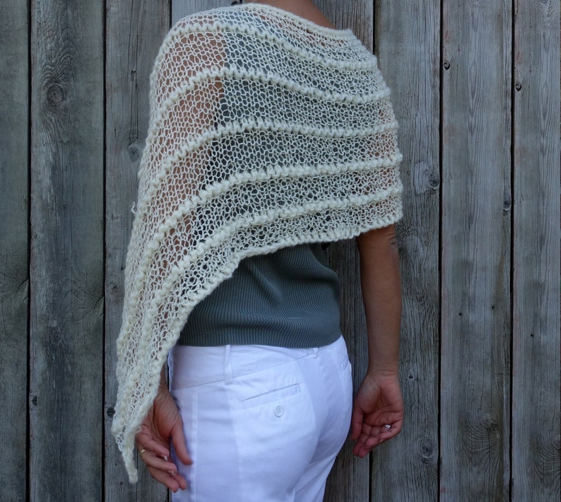 May include: A white crocheted shawl with a lacy pattern. The shawl is draped over a person's shoulders and is worn with a gray shirt and white pants.