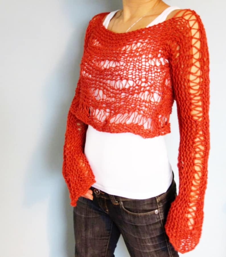 May include: A red crocheted sweater with a loose, open knit pattern. The sweater has long sleeves and a cropped length. The sweater is worn over a white shirt and blue jeans.