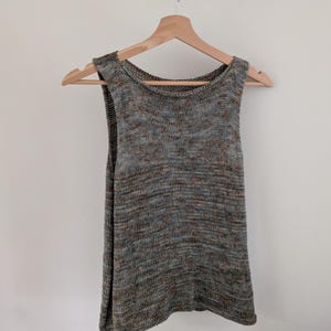 May include: A sleeveless knit top in a neutral color palette of gray, brown, and blue, hanging on a wooden hanger. The top has a scoop neckline and a slightly flared shape.