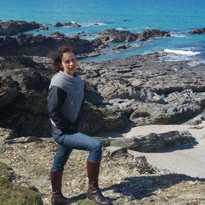 Puede incluir: Una mujer con una bufanda de punto gris y pantalones vaqueros azules se encuentra en un acantilado rocoso con vistas a un oc&eacute;ano azul. Lleva botas de cuero marr&oacute;n.