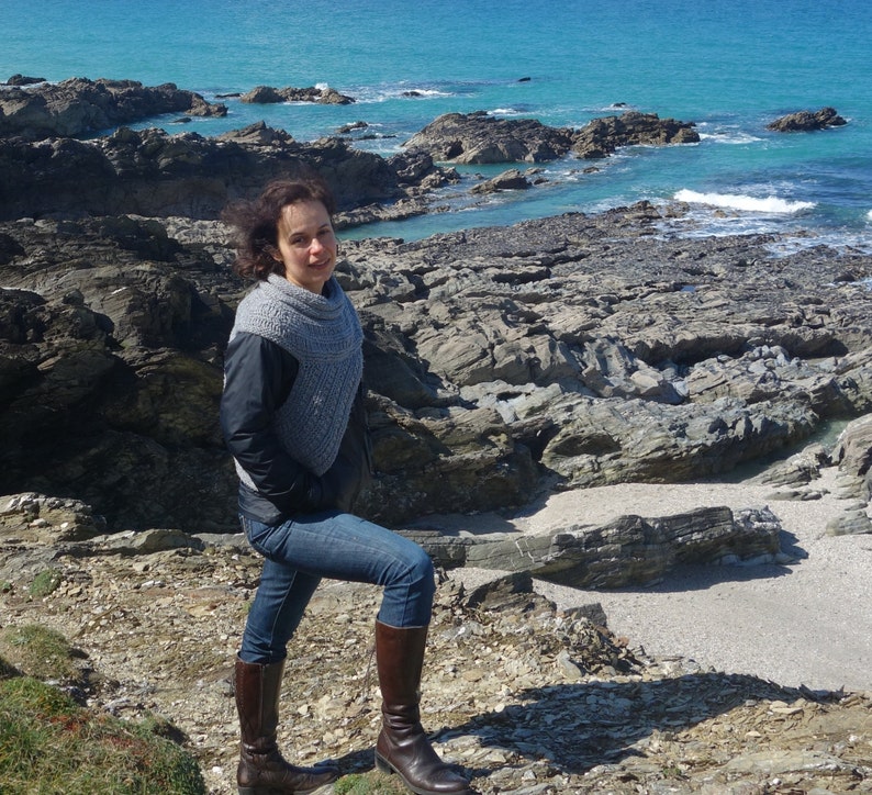 Puede incluir: Una mujer con una bufanda de punto gris y pantalones vaqueros azules se encuentra en un acantilado rocoso con vistas a un oc&eacute;ano azul. Lleva botas de cuero marr&oacute;n.