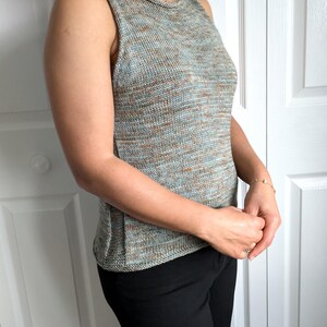 May include: A sleeveless, knitted top in a blend of blue, brown, and gray yarns. The top has a round neckline and a textured appearance. The person is wearing black pants. The top appears to be handmade.