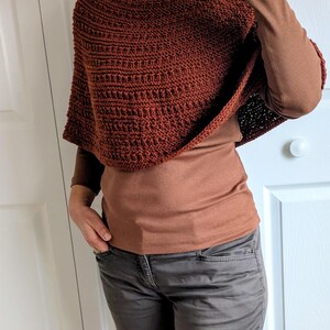 Pode incluir: Um poncho de cor ferrugem, feito &agrave; m&atilde;o em croch&eacute;, com um padr&atilde;o de pontos canelados texturados. A pe&ccedil;a &eacute; usada sobre uma camisola castanha de manga comprida e combinada com cal&ccedil;as cinzentas escuras. O poncho tem um decote redondo.