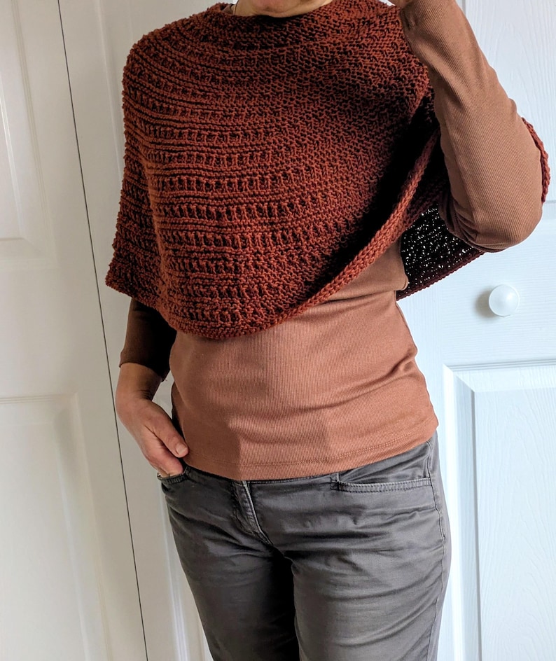 Pode incluir: Um poncho de cor ferrugem, feito &agrave; m&atilde;o em croch&eacute;, com um padr&atilde;o de pontos canelados texturados. A pe&ccedil;a &eacute; usada sobre uma camisola castanha de manga comprida e combinada com cal&ccedil;as cinzentas escuras. O poncho tem um decote redondo.