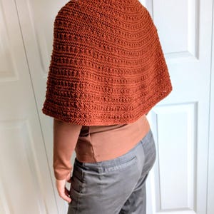 Pode incluir: Um poncho de cor ferrugem, feito &agrave; m&atilde;o em croch&eacute;, com um padr&atilde;o de pontos canelados texturizados. A pe&ccedil;a &eacute; usada sobre uma camisa castanha de manga comprida e cal&ccedil;as cinzentas, cobrindo os ombros e a parte superior das costas.