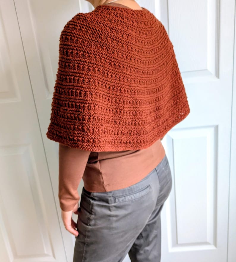 Pode incluir: Um poncho de cor ferrugem, feito &agrave; m&atilde;o em croch&eacute;, com um padr&atilde;o de pontos canelados texturizados. A pe&ccedil;a &eacute; usada sobre uma camisa castanha de manga comprida e cal&ccedil;as cinzentas, cobrindo os ombros e a parte superior das costas.