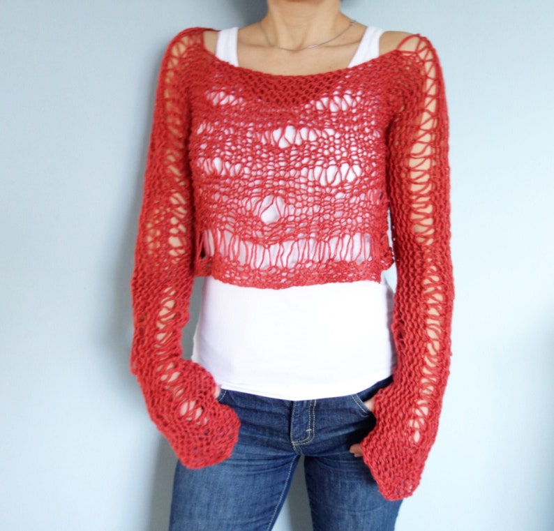 May include: A red crocheted sweater with a loose, open knit. The sweater has long sleeves and a cropped length. The sweater is worn over a white tank top and blue jeans.