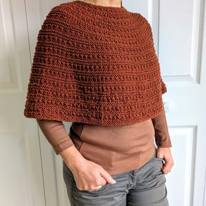 Pode incluir: Um poncho tricotado &agrave; m&atilde;o na cor ferrugem com um padr&atilde;o de pontos texturizado e repetitivo. A pe&ccedil;a tem um decote alto e arredondado e uma silhueta curta tipo capa que cai sobre os ombros, usada sobre uma camisa castanha de manga comprida.