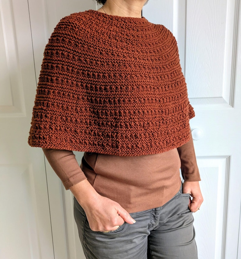 Pode incluir: Um poncho tricotado &agrave; m&atilde;o na cor ferrugem com um padr&atilde;o de pontos texturizado e repetitivo. A pe&ccedil;a tem um decote alto e arredondado e uma silhueta curta tipo capa que cai sobre os ombros, usada sobre uma camisa castanha de manga comprida.