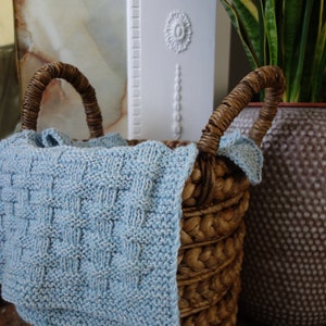 May include: A woven wicker basket with a light blue knitted blanket inside. The basket has two handles and is sitting on a wooden surface.