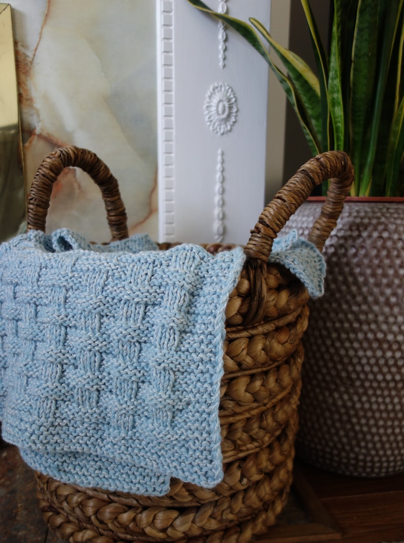 May include: A woven wicker basket with a light blue knitted blanket inside. The basket has two handles and is sitting on a wooden surface.
