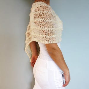 May include: A white crocheted shawl with a delicate openwork pattern. The shawl is draped over the shoulders of a person wearing a white top and white pants.