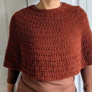 Pode incluir: Um poncho tricotado &agrave; m&atilde;o na cor ferrugem, com um padr&atilde;o de pontos texturizados que apresenta filas horizontais de relevos e detalhes abertos. A pe&ccedil;a tem um decote redondo e uma silhueta curta tipo capa que cai sobre os ombros.