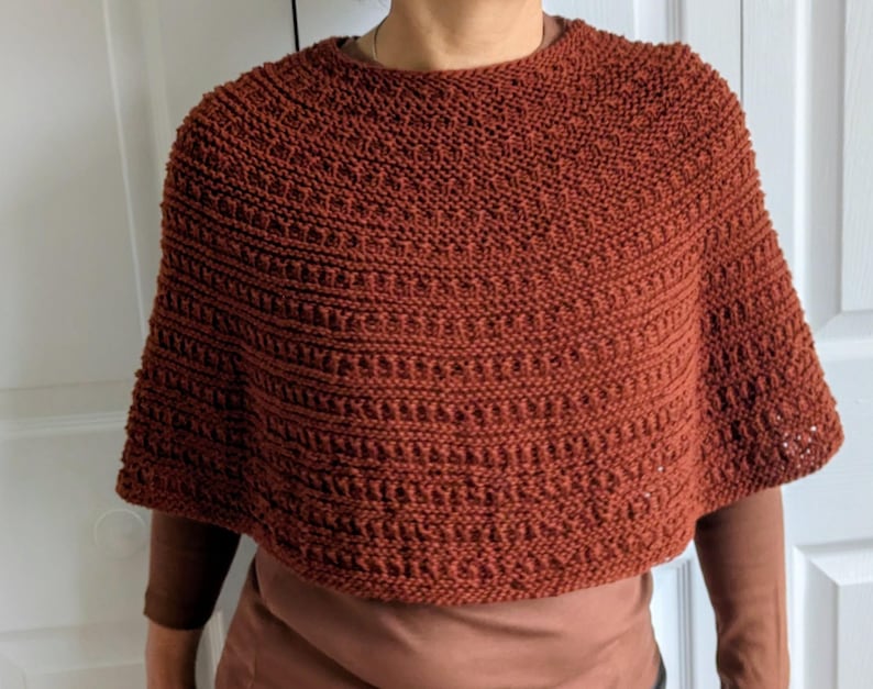 Pode incluir: Um poncho tricotado &agrave; m&atilde;o na cor ferrugem, com um padr&atilde;o de pontos texturizados que apresenta filas horizontais de relevos e detalhes abertos. A pe&ccedil;a tem um decote redondo e uma silhueta curta tipo capa que cai sobre os ombros.