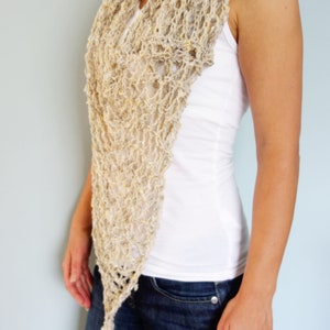May include: A beige crochet scarf with a lacy pattern. The scarf is draped around the neck of a person wearing a white tank top and blue jeans.