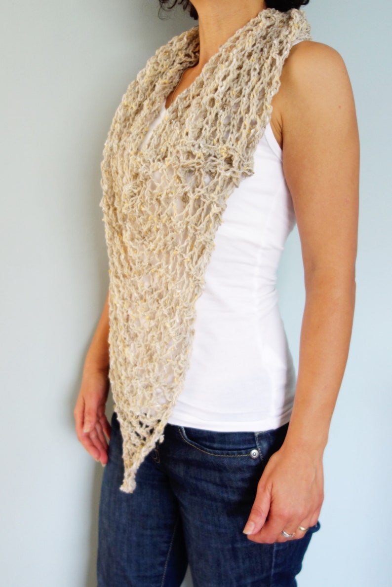 May include: A beige crochet scarf with a lacy pattern. The scarf is draped around the neck of a person wearing a white tank top and blue jeans.