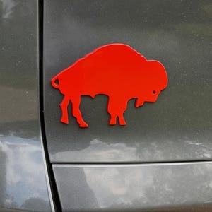 May include: A bright red, die-cut buffalo emblem affixed to the rear of a gray vehicle. The emblem is a silhouette of the animal, showcasing its distinctive shape and features. The image is a close-up, highlighting the emblem's color and design.