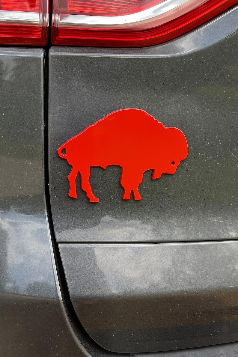 May include: A bright red, die-cut buffalo emblem affixed to the rear of a gray vehicle. The emblem is a silhouette of the animal, showcasing its distinctive shape and features. The image is a close-up, highlighting the emblem's color and design.