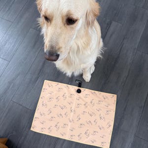May include: A Golden Retriever sits near a peach-colored mat with a repeating pattern of dog silhouettes. The mat has a black handle and button closure. The dog has light-colored fur and is looking down.