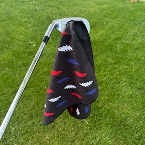 May include: A black golf towel with a pattern of red, white, and blue feather-like shapes is draped over a golf club. The towel is made of a textured material and is hanging on a golf club on a green grassy lawn.