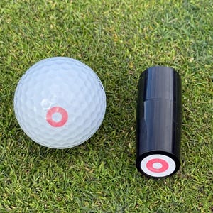 May include: A white golf ball with a red circle on it and a black golf ball marker with a red circle on it.