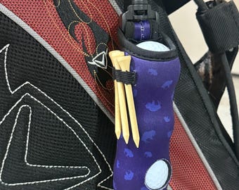 May include: A purple golf ball holder with a blue pattern, holding a white golf ball and wooden tees. The holder is attached to a black golf bag with red accents. The holder has a clip for easy attachment.
