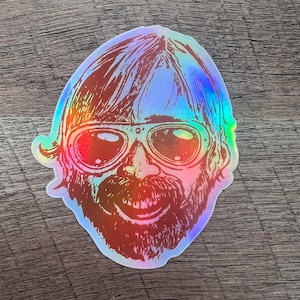 May include: Holographic sticker featuring a smiling face with sunglasses, a beard, and long hair. The sticker has a rainbow sheen, reflecting various colors. The design is a stylized illustration.