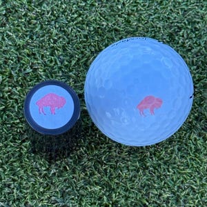May include: A white golf ball with a red buffalo logo and a black golf ball marker with a blue background and a red buffalo logo.