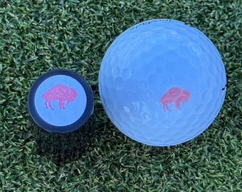 May include: A white golf ball with a red buffalo logo and a black golf ball marker with a blue background and a red buffalo logo.
